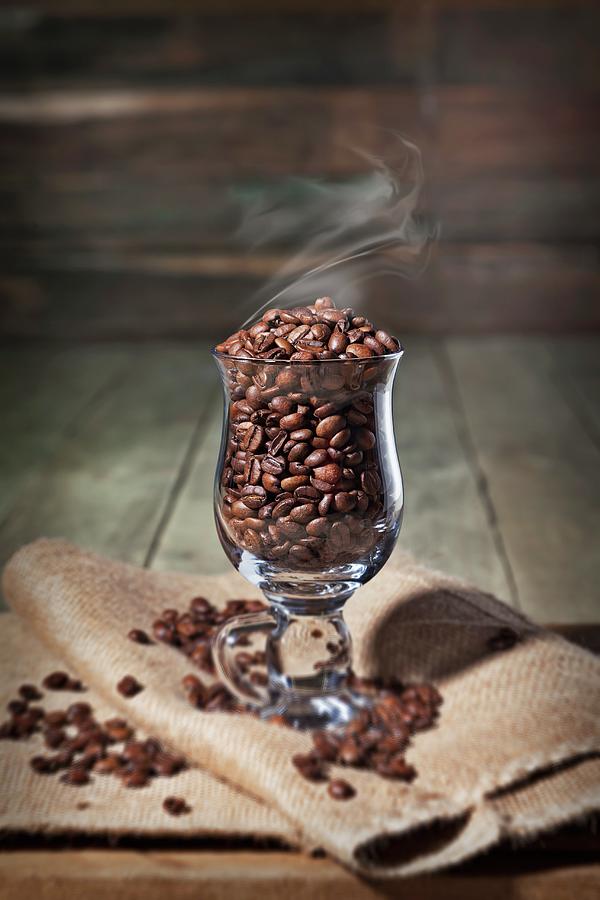 A Glass Coffee Cup With Steaming Coffee Beans On A Folded Burlap Bag Photograph by George Crudo