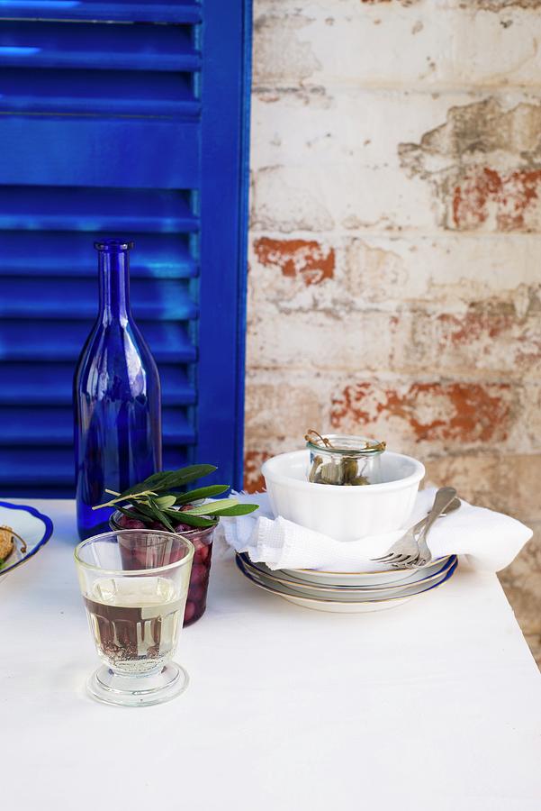 A Glass Of White Wine On A Table With A White Tablecloth, Stacked Crockery, Kalamata Olives And A Wine Bottle In Front Of A Royal Blue Wooden Shutter Photograph by Great Stock!