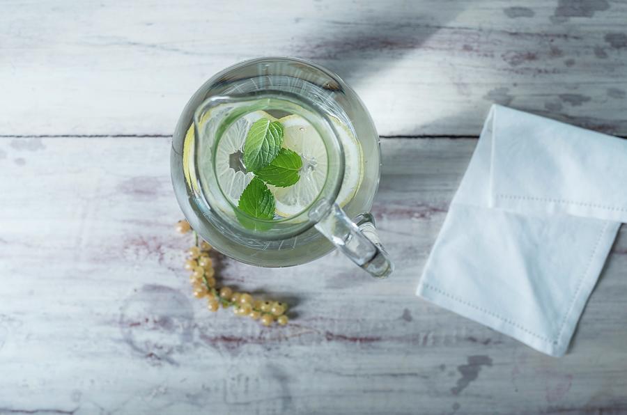 A Glass Water Jug With Lemon And Mint On A White Wooden Surface Next To Whitecurrants And A Fabric Napkin Photograph by Angelika Grossmann