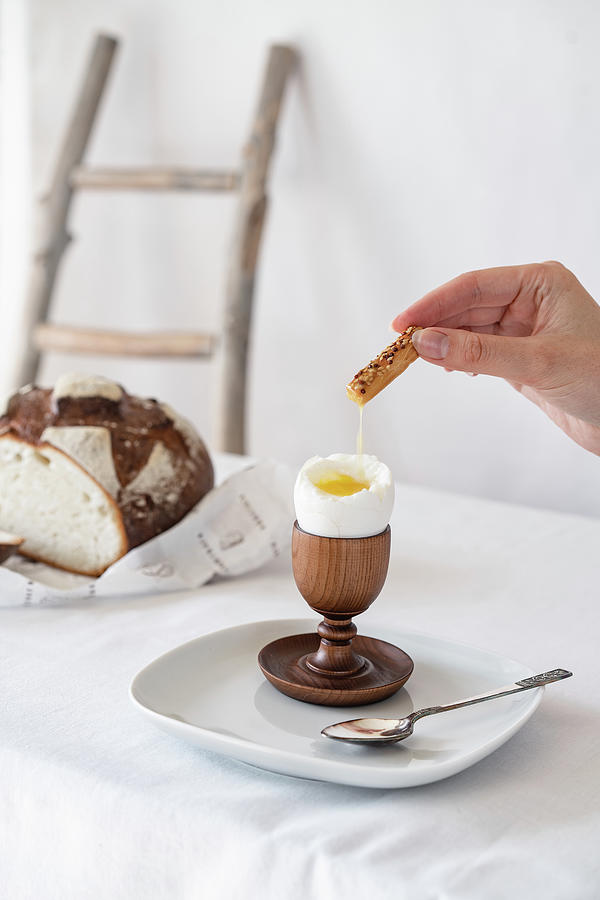 Easter Photograph - A Hand Soaks A Cracker Into The Yolk Of A Broken Boiled Egg On A Wooden Stand On A Table With A White Tablecloth by Cavan Images