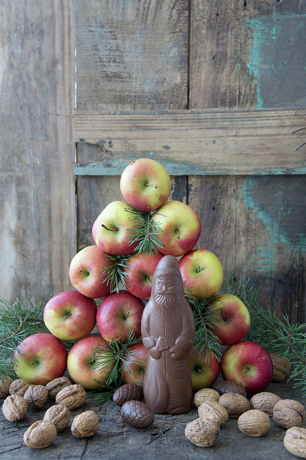 A Homemade Chocolate Santa Claus In Front Of A Pyramid Of Apples And Pine Branches Photograph by Martina Schindler