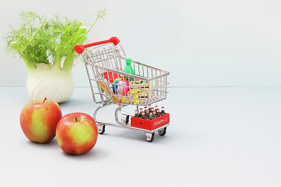 A Mini Shopping Trolley Filled With Toy Foodstuffs Next To Apples And A Bulb Of Fennel Photograph by Misha Vetter