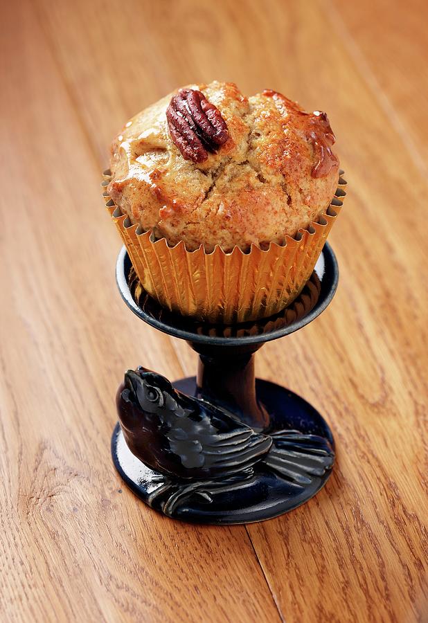 A Pecan And Maple syrup Muffin In A Gold Muffin Case On A Small Blue Stand With A Ceramic Bird All Sitting On A Wooden Surface Photograph by Stuart Macgregor