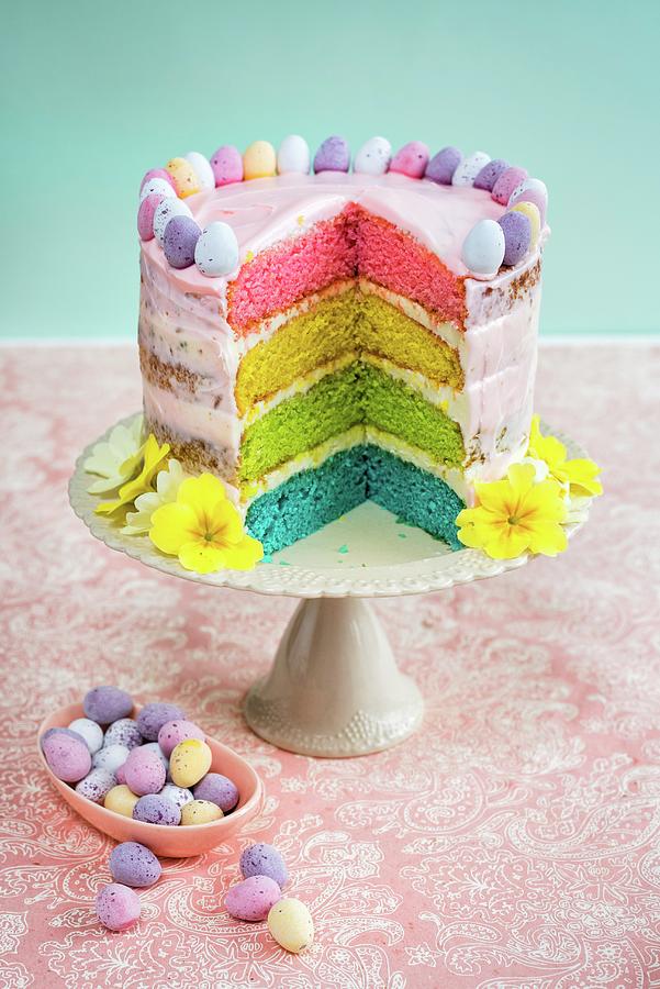 A Rainbow Cake Decorated With Mini Chocolate Eggs And Flowers For Easter with A Slice Cut Out Photograph by Lucy Parissi