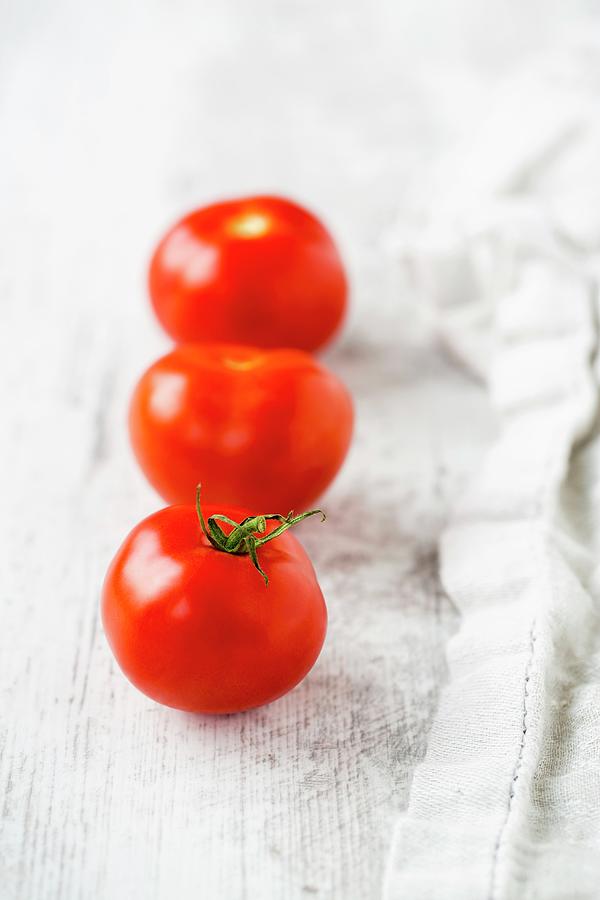 A Row Of Three Tomatoes Next To A Tea Towel On A White Wooden Surface Photograph by Sandra Krimshandl-tauscher