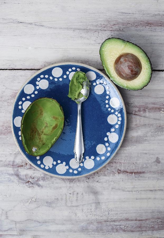 A Scooped Out Avocado Half On A Blue Clay Plate With A Spoon Next To Half An Avocado Photograph by Angelika Grossmann