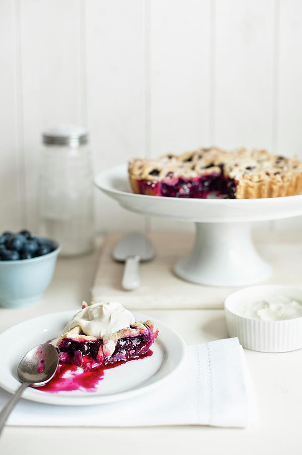 A Slice Of Blueberry Pie With Cream On A Plate With The Rest Of The Pie On A Cake Stand In The Background Photograph by Magdalena Hendey