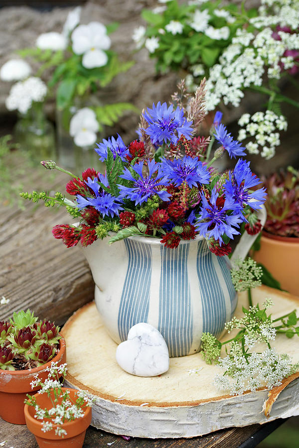 A Small Bouquet Of Cornflowers And Crimson Clovers In A Pitcher On A Disc Of Birch Wood, A Heart Made Of Howlite Crystal Photograph by Angelica Linnhoff