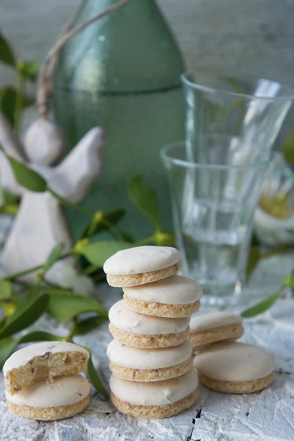 A Stack Of Anise Biscuits With Shot Glasses, A Green Bottle And An Angel Figurine In The Background Photograph by Martina Schindler