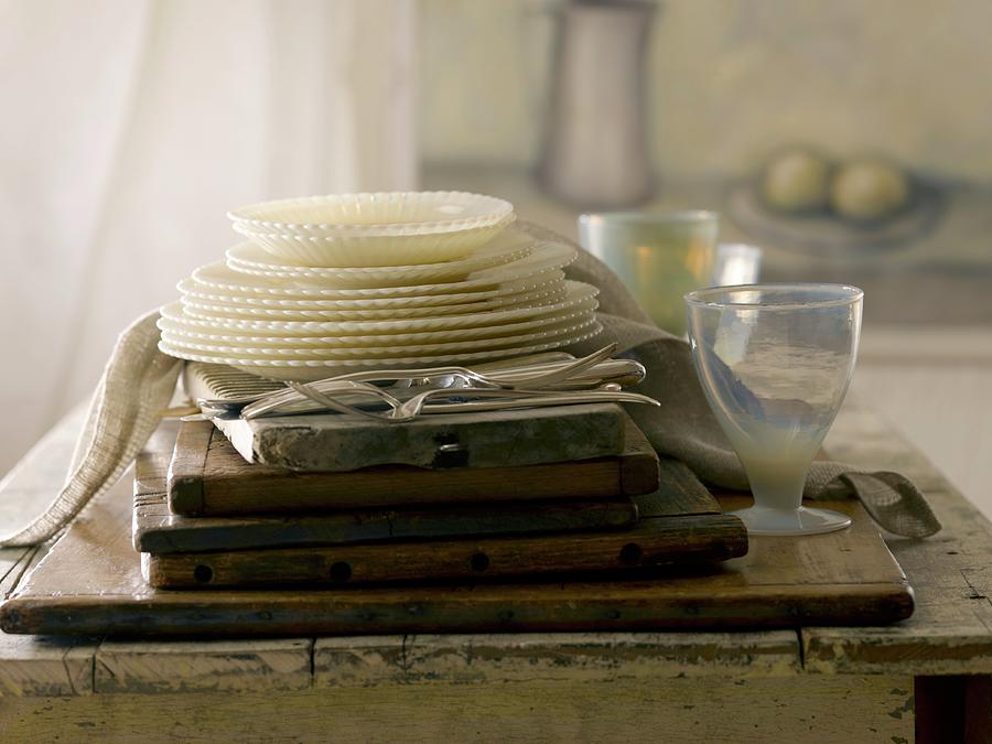 A Stack Of Vintage Plates, Glassware And Cutlery In Front Of A Painting And A Curtain Photograph by Laurie Proffitt