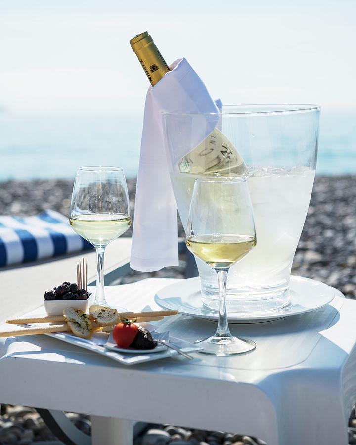 A Table Laid For Two At The Beach, With Appetisers, Glasses Of White Wine And A Bottle Of Wine In A Chiller Photograph by Lanneretonne, Anthony