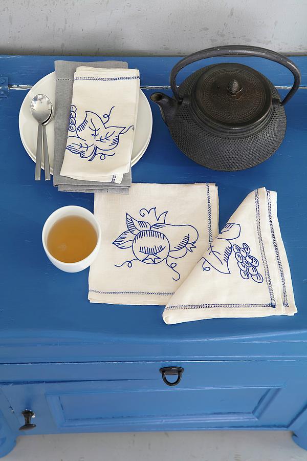 A Teapot, A Tea Bowl And Linen Napkins Embroidered With Various Motifs On A Blue Chest Of Drawers Photograph by Jalag / Olaf Szczepaniak
