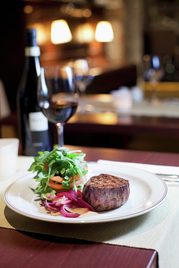A View On The Interior Of The Restaurant, A Table With Wine, A Dish Of Sirloin Steak With Red Onions, Tomatoes And Rocket Leaves Photograph by Jan Prerovsky