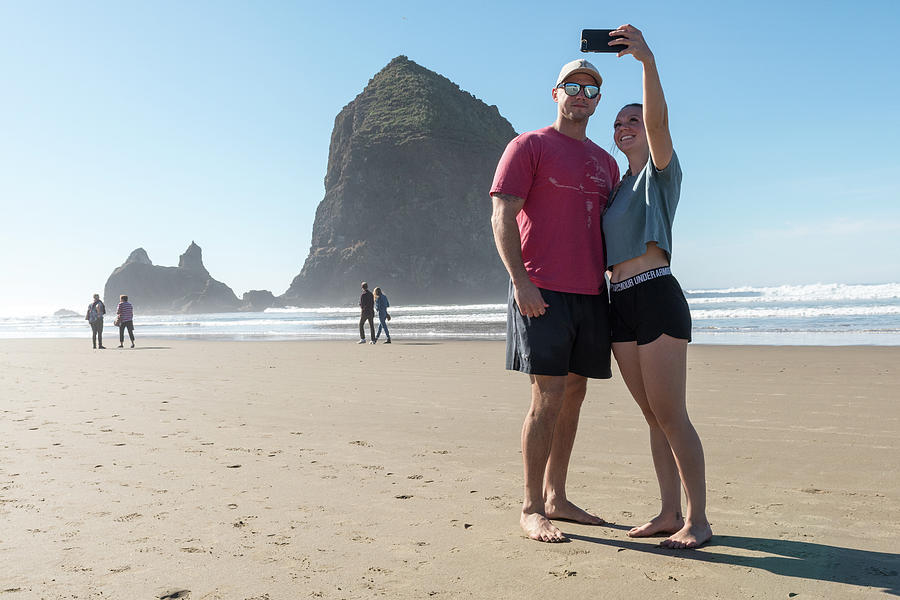 A Young Couple Of Tourists Make Selfies In Front Of The Haystackcannon Beach, Oregon, Usa - October Photograph