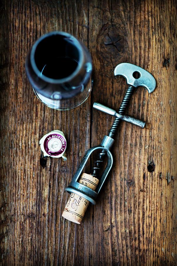 An Antique Corkscrew With A Cork, And A Glass Of Red Wine On A Wooden Surface Photograph by Jamie Watson