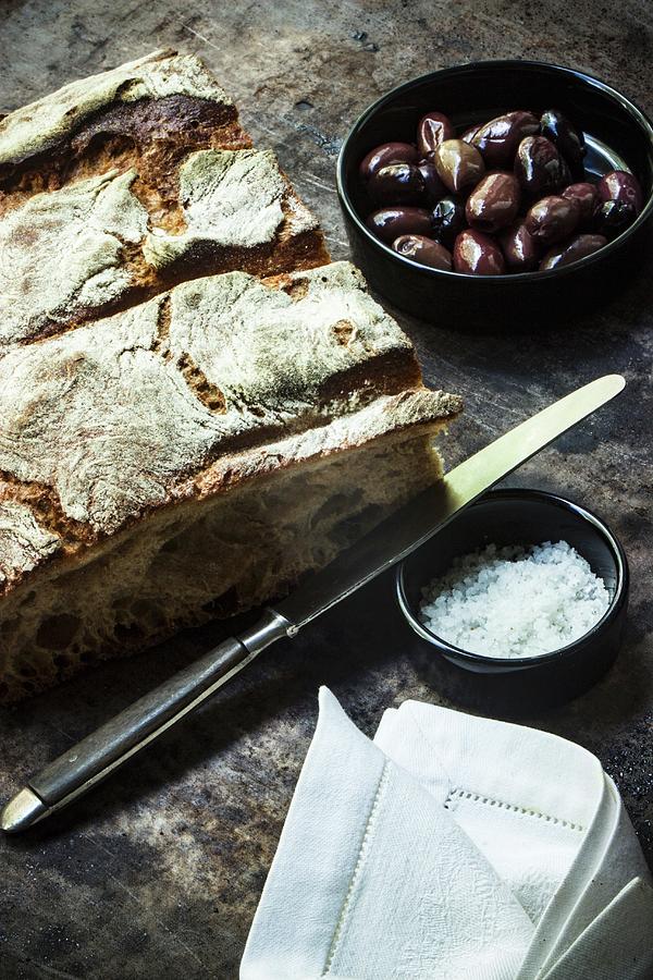 An Arrangement Of Crusty Bread, Olives, Sea Salt, A Knife And A Fabric Napkin Photograph by Charlotte Von Elm