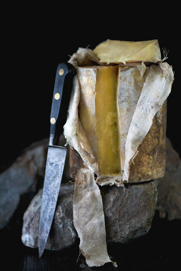An Entire Mature Cave Aged Cheddar Cheese With Its Cloth Rolled Back And A Slice Removed On A Dark Rock With A Vintage Knife Photograph by Jamie Watson