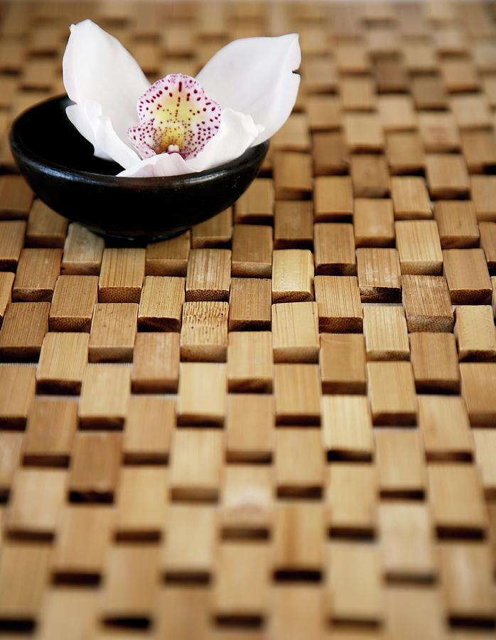 An Orchid Flower In A Little Black Bowl On A Bamboo-wood Mat Photograph by Great Stock!