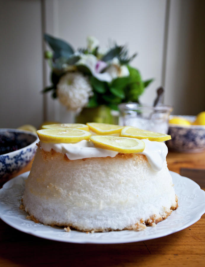 Angel Food Cake With Whipped Cream And Lemon Slices, With Flowers, A Jar Of Whipped Cream, And Bowl Of Lemons In The Background Photograph by Ryla Campbell