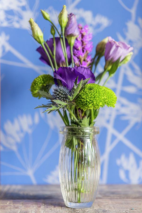 Arrangement Of Purple Flowers In Glass Vase In Front Of Wall With Floral Pattern Photograph by Stuart Cox