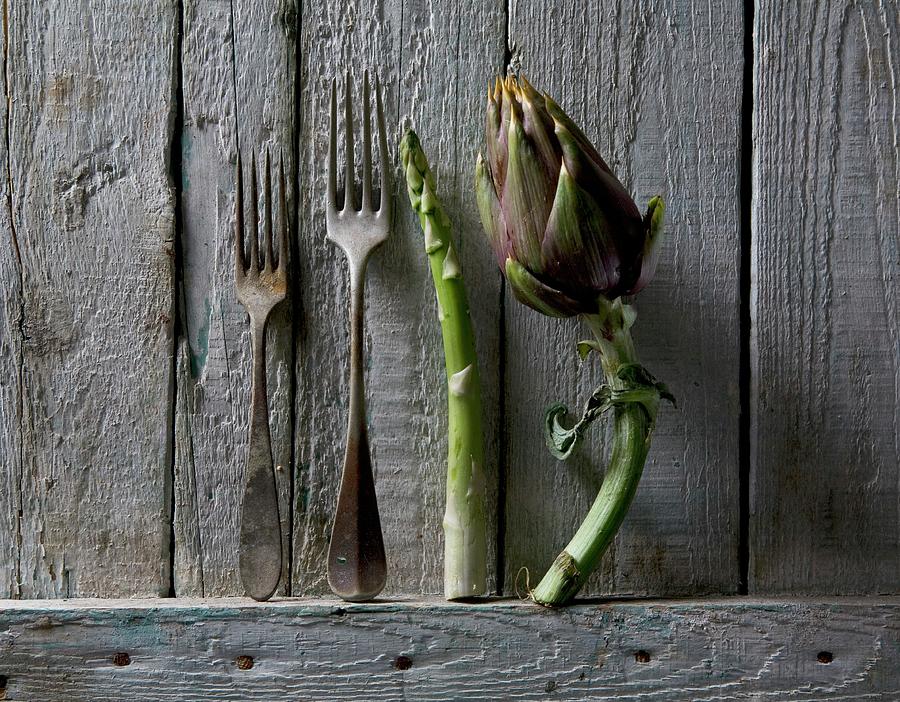 Artichoke, Asparagus And Rusty Forks Against A Rustic Wooden Wall Photograph by Blueberrystudio