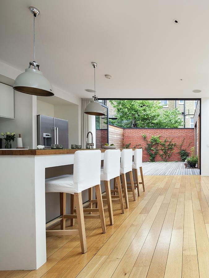 Bar Stools With White Loose Covers At Kitchen Counter In Open-plan Designer Kitchen; Open Folding Terrace Doors In Background Photograph by Simon Maxwell Photography