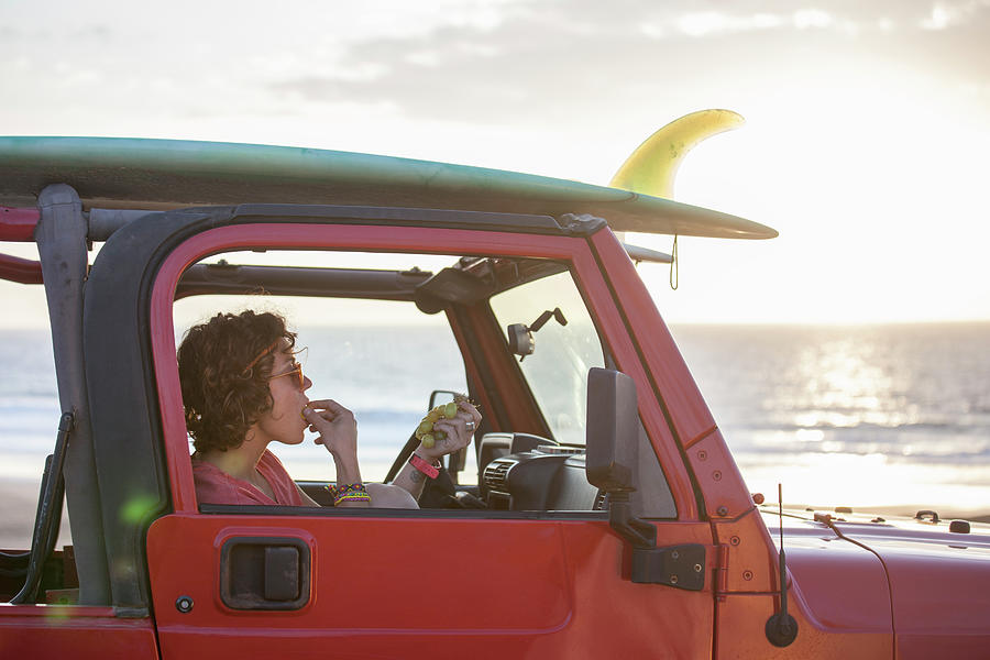 Sunset Photograph - Beautiful Girl In Her Jeep Eating Grapes With Surfboard On The Roof by Cavan Images
