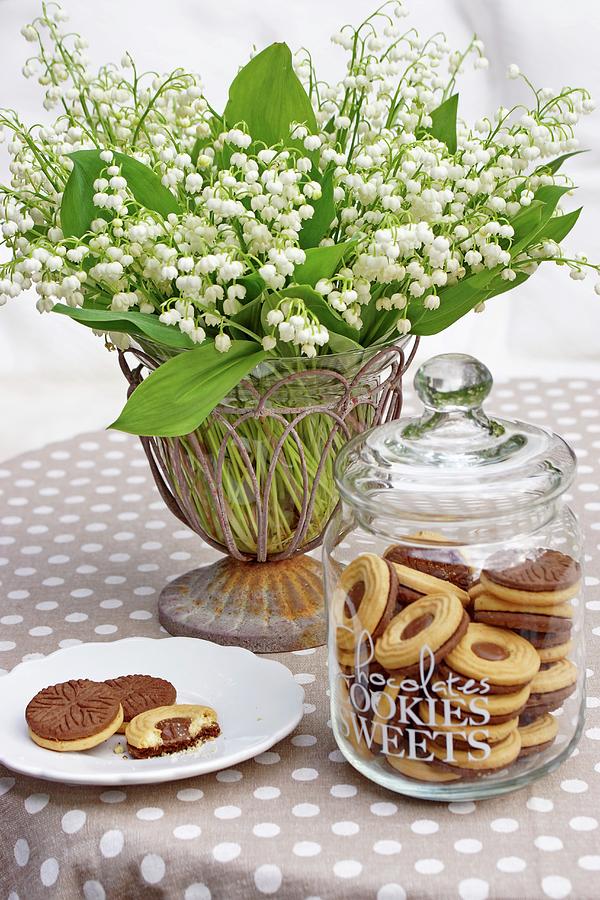 Biscuits In Glass Jar And On Plate In Front Of Vase Of Lily-of-the-valley On Brown Tablecloth With White Polka Dots Photograph by Angelica Linnhoff