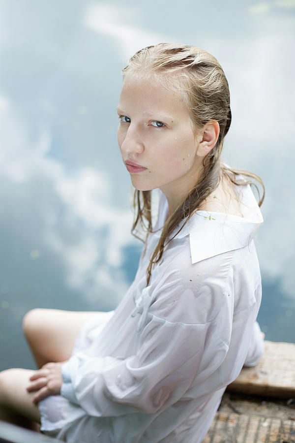 Blonde Teenage Girl In White Wet Shirt Is Sitting By The Blue Water