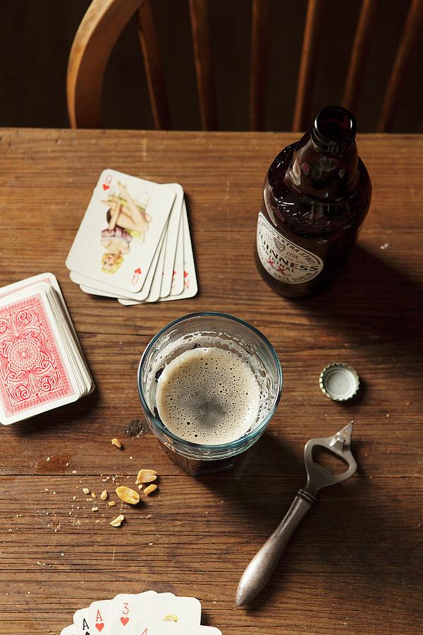 Bottle Of Guinness Poured Into A Large Glass On A Wooden Table Surrounded With Bottle Top, Metal Bottle Opener, Nuts And Playing Cards Photograph by Stacy Grant