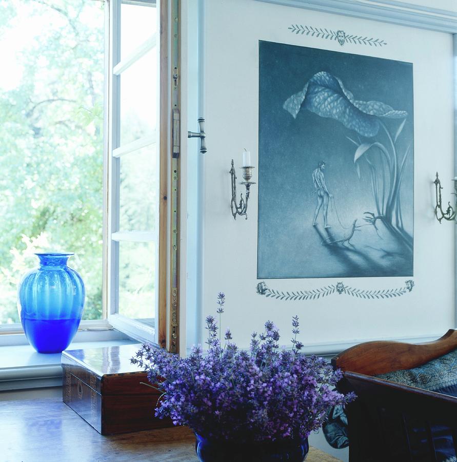 Bowl Of Purple Flowers On Table In Front Of Window With Blue Glass Vase On Windowsill; Painting In Shades Of Blue To One Side Photograph by Inge Ofenstein