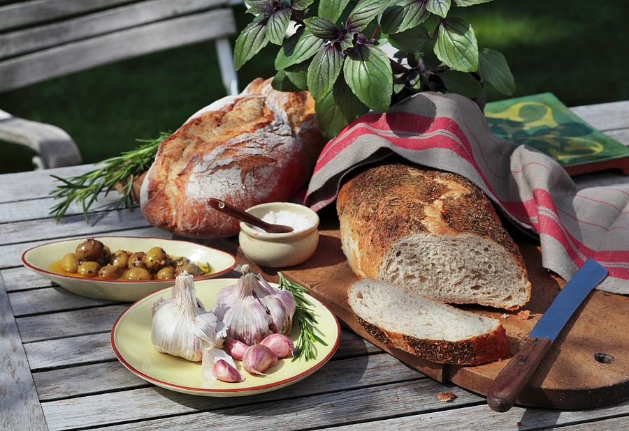 Bread, Fresh Herbs, Garlic, Olives And Salt On A Garden Table Photograph by Peter Garten