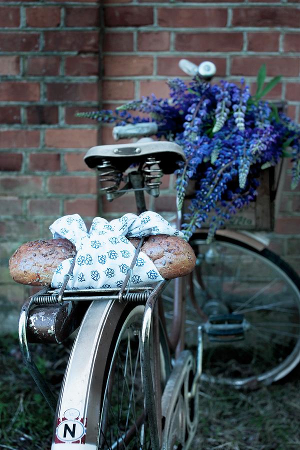 Bread Wrapped In Blue And White Cloth On Bicycle Luggage Rack With Bouquet Of Lupins In Crate On Handlebars In Front Of Brick Wall Photograph by Annette Nordstrom