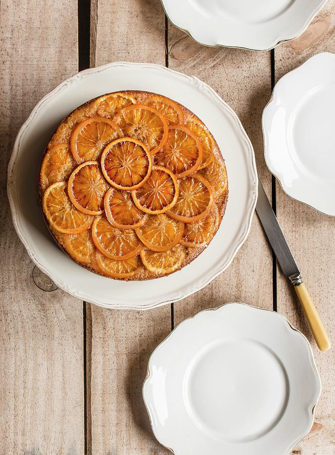 Cake Topped With Orange Slices On White Cake Stand, With White Plates And Wooden Background Photograph by Miriam Garcia