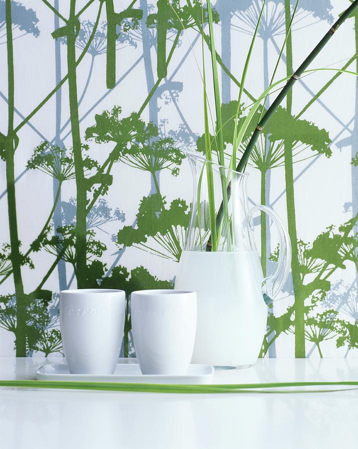 Carafe And White Ceramic Beakers In Front Of Herb-patterned Wallpaper