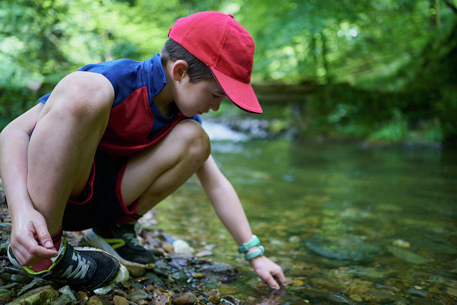 Summer Photograph - Child With Red Hat By The River Bank Playing With Water And Stones In The Forest by Cavan Images