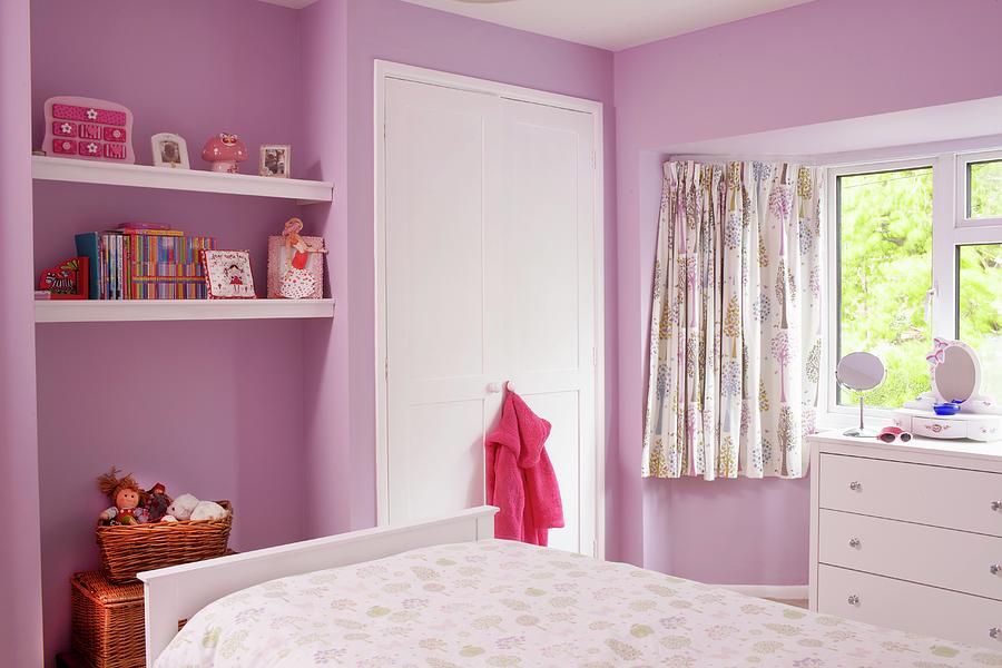 Childs Bedroom With Pink-painted Walls, Floating Shelves In Niche, White Fitted Wardrobe And Chest Of Drawers Below Window Photograph by Tim Imri