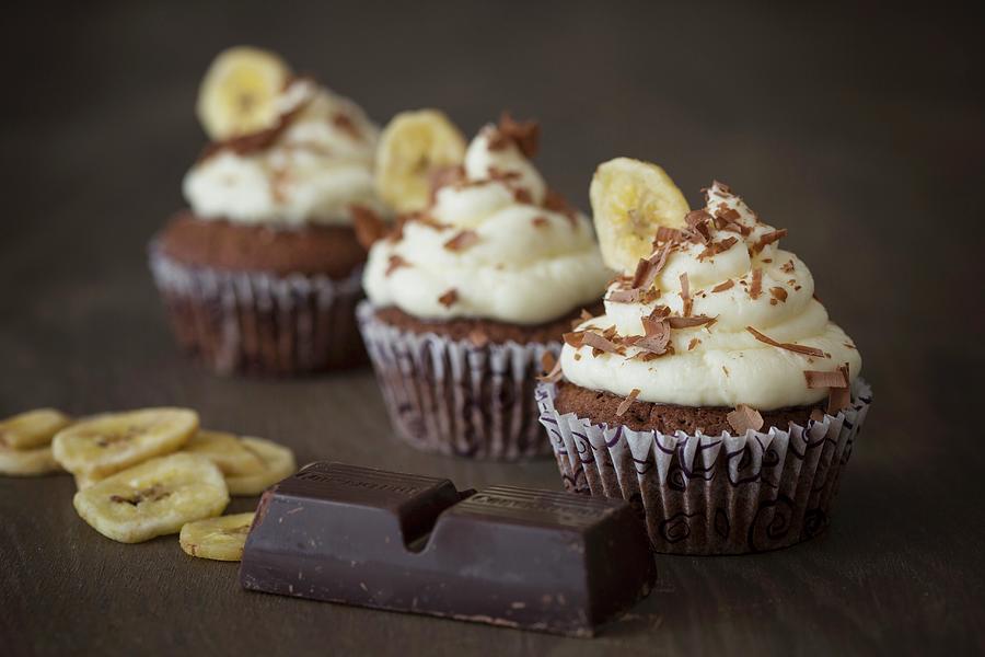 Chocolate Cupcakes With A Banana Topping With Cooking Chocolate And Banana Chips Next To It Photograph by Jan Wischnewski