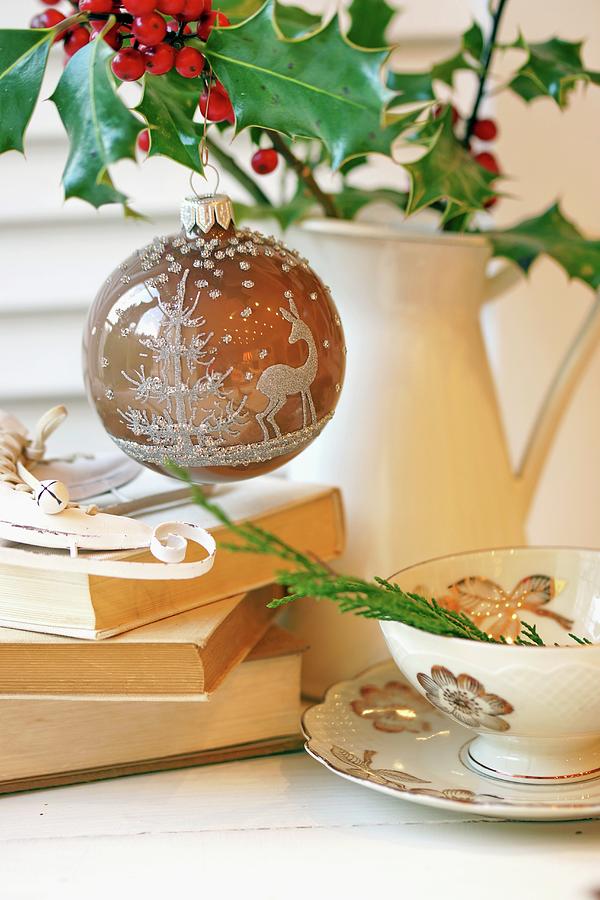 Christmas Bauble With Forest Motif Hanging From Holly In Jug Next To Stack Of Books And Teacup Photograph by Angelica Linnhoff