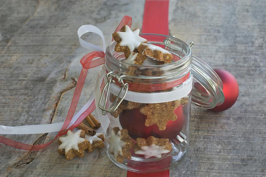 Cinnamon Stars In A Jar In Front Of Christmas Tree Baubles And A Ribbon Photograph by Achim Sass