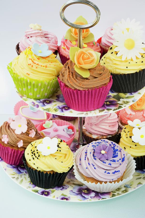 Close Up Of A Selction Of Iced Purple, Yellow And Pink Cup Cakes Decorated With Icing Flowers On A Purple Pansy Flowered Cake Stand And A White Background Photograph by Burgess, Linda