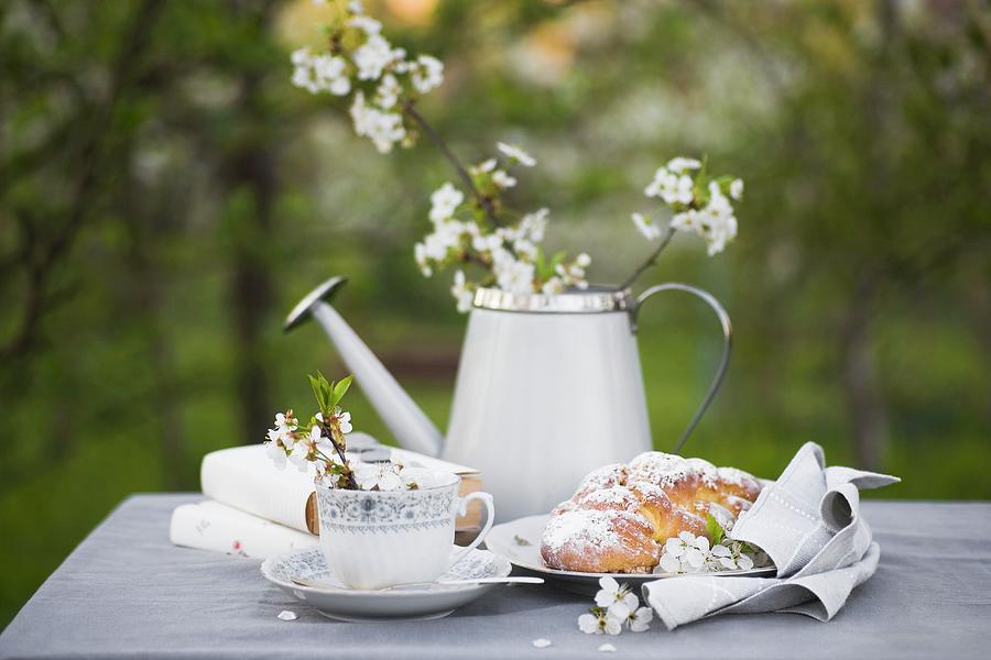 Coffee Cup, Books, Plaited Bun, And Flowers In Watering Can On Grey Tablecloth Photograph by Alicja Koll