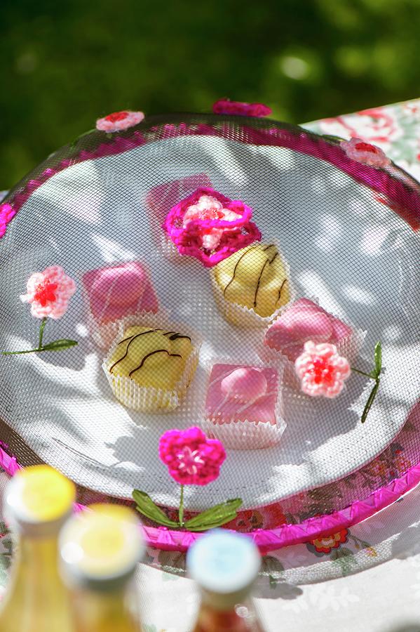Colourful Petit Fours Under Mesh Cake Cover Decorated With Crocheted Flowers On Garden Table Photograph by Winfried Heinze