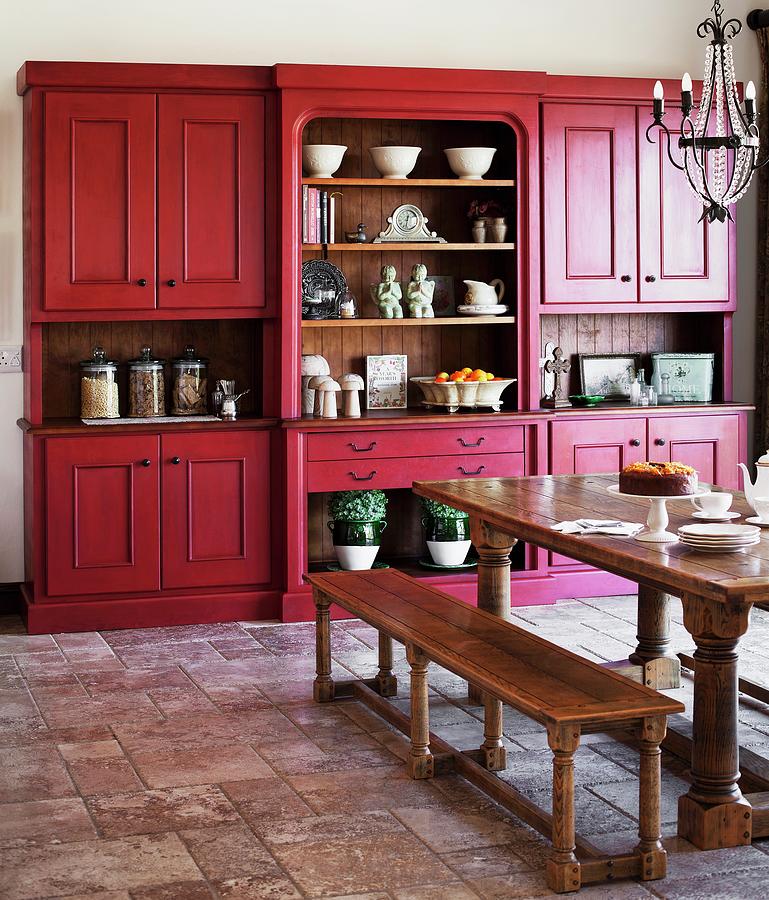 Country-house Style Kitchen-dining Room With Heavy Wooden Table And Benches Below Wrought Iron Chandelier; Matt Red Kitchen Dresser In Background Photograph by Great Stock!