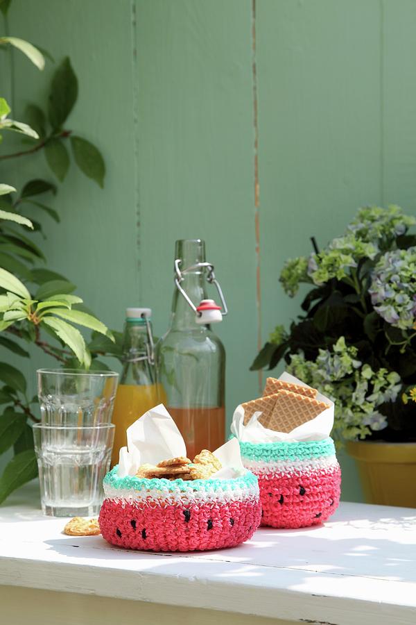 Crocheted Watermelon Baskets Of Biscuits Next To Glasses And Swing-top Bottles On Garden Table Photograph by Thordis Rggeberg