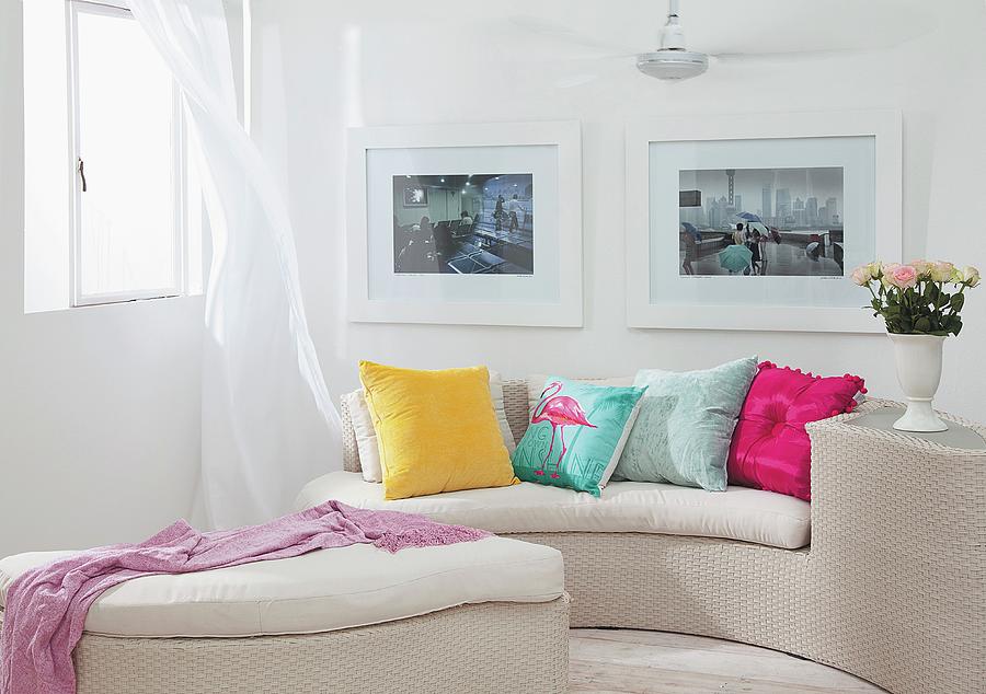 Curved, Pale Wicker Sofa And Matching Ottoman With Colourful Scatter Cushions On Seat Cushions In Corner Below Framed Pictures On Wall Photograph by Great Stock!