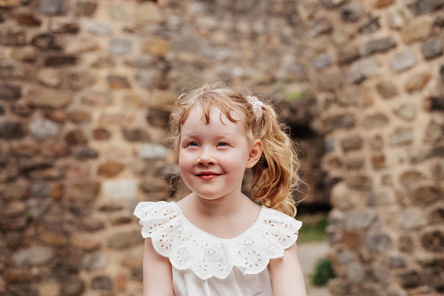 Architecture Photograph - Cute Smiling Girl With Blond Hair Looking Away While Sitting Against Old Brick Wall by Cavan Images