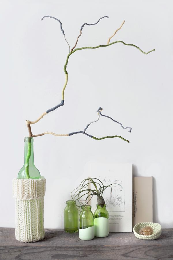 Decorative Arrangement Of Branch Covered In Wool Remnants Of Various Colours, Glass Vase With Knitted Cover, Three Small Bottles, Air Plant And Books Photograph by Sabine Lscher
