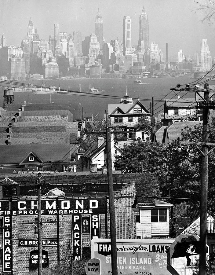 New York City Photograph - Distant view of lower Manhattan and ferry docks as seen w. the aid of a telephoto lens over the rooftops of buildings in Staten Island. by Andreas Feininger