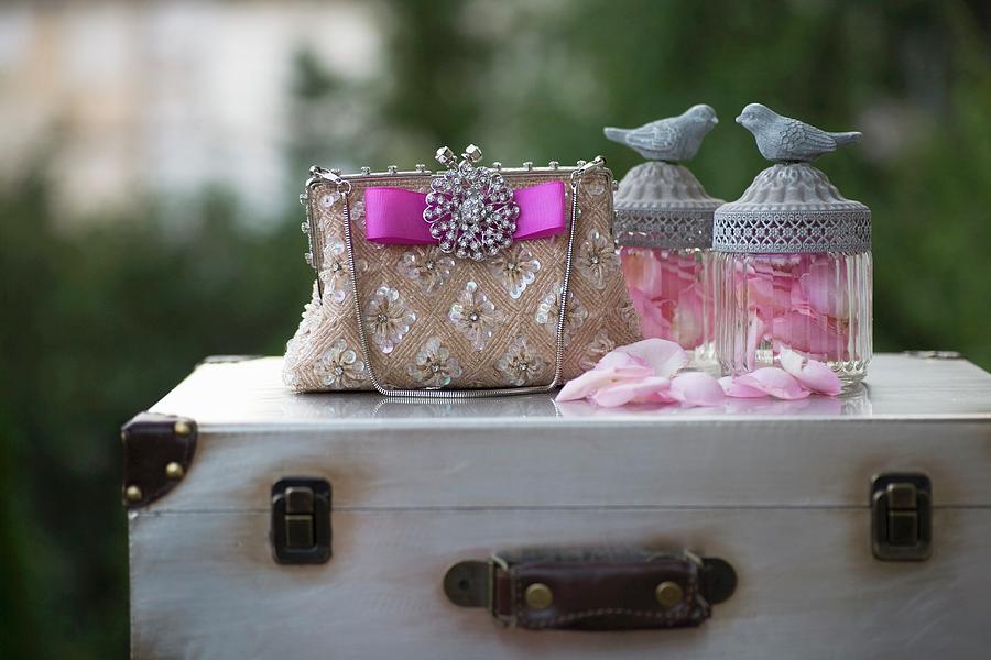 Elegant, Sequinned Handbag With Brooch And Pink Ribbon And Glass Jars Of Rose Petals With Bird Figurines On Lids On Top Of Vintage Suitcase Photograph by Alicja Koll